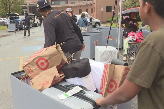 a student pushes a large wheeled tote of items for his dorm room with the help of a move-in volunteer.