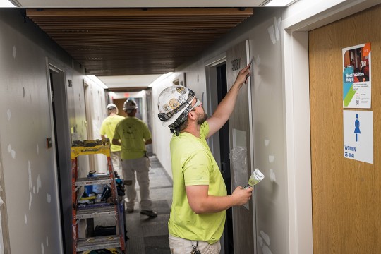 a man in a lime green shirt and hard hair adds spackle to a wall in an R I T dorm hallway.