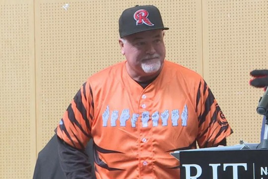 a man stands in front of a podium wearing a black and orange tiger stripe jersey that reads Rochester in finger spelling across the front.