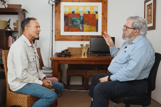 two men sit facing each other in chairs in a home office.