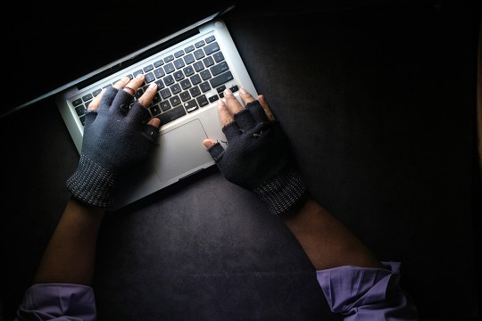 gloved hands type on a laptop sitting in a dark room.