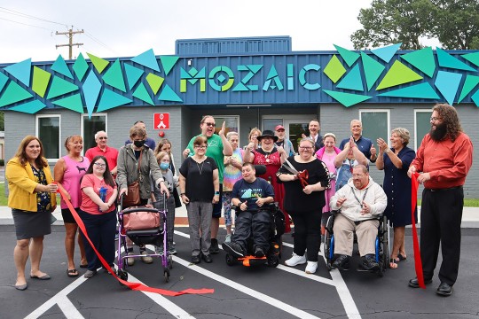 'people with many different abilities are shown at a ribbon cutting ceremony in front of a building with a sign that says MOZAIC'