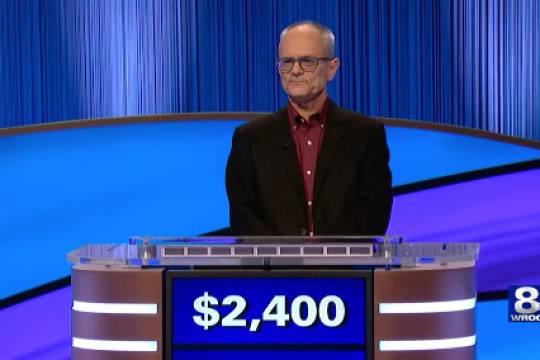 David Bond appears on the set of Jeopardy. The sign in front of him reads his current winnings, twenty four hindred dollars.