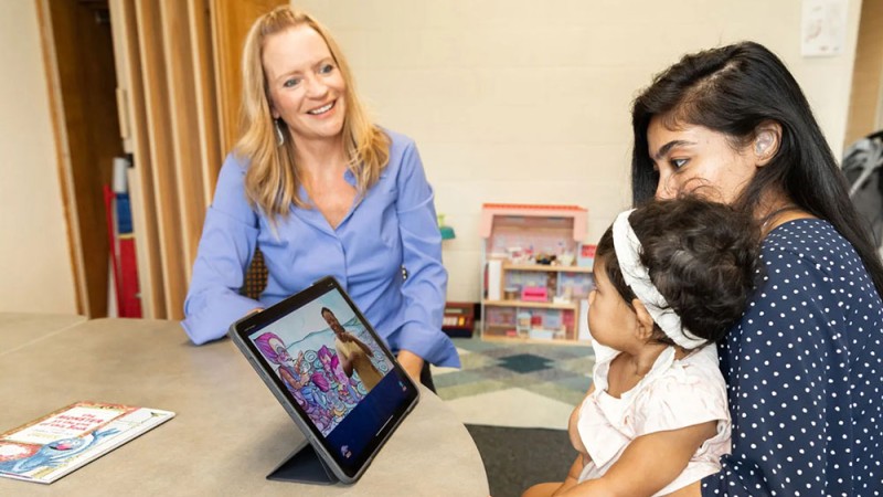 RIT professor Rain Bosworth wears a blue blouse and sits at a table with a mother who has a small child sitting on her lap. They are looking at a computer tablet.