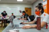 students wearing masks sitting at a long table working on laptops.