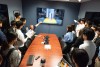 several students gather around a long table watching a screen placed at one end.