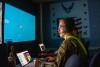 An R O T C cadet sits at a control panel looking at a large screen in front of them.