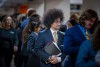 Young man in a navy suit and yellow tie stands in a crowded hallway at a career fair, holding a notebook and looking ahead with a confident expression.