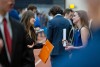 Two women in business attire have a conversation at a career fair, one holding an orange R I T folder while the other holds a rolled-up document.