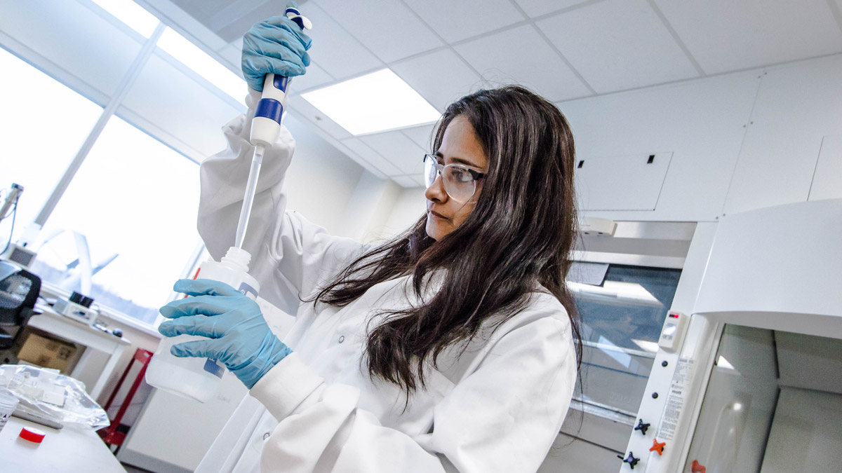 A student with long brown hair in a lab uses a pipette to put liquid into a bottle.