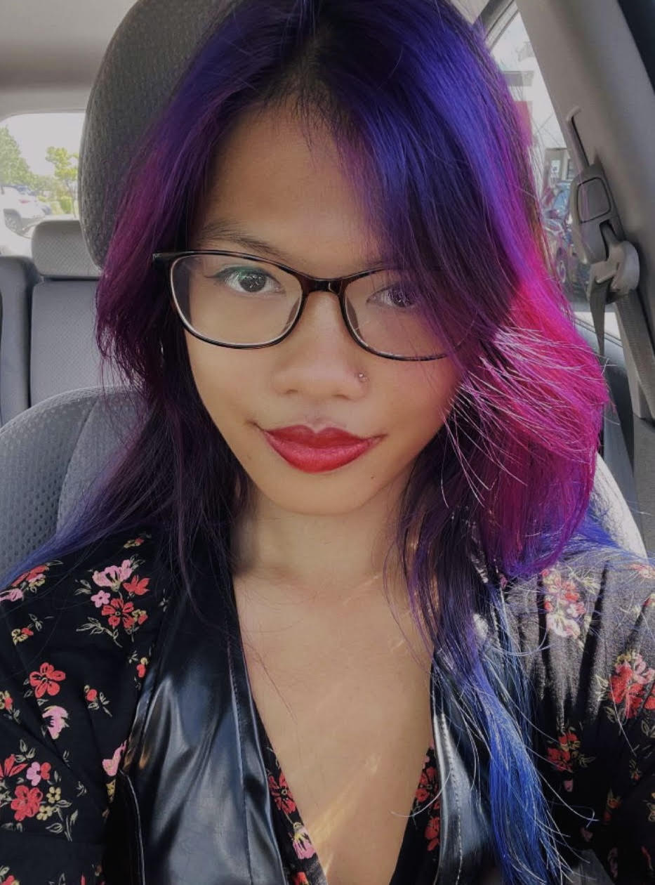 An Asian woman with purple and pink long hair, nose ring, glasses, and red lipstick looks at the camera from the front sear of their car