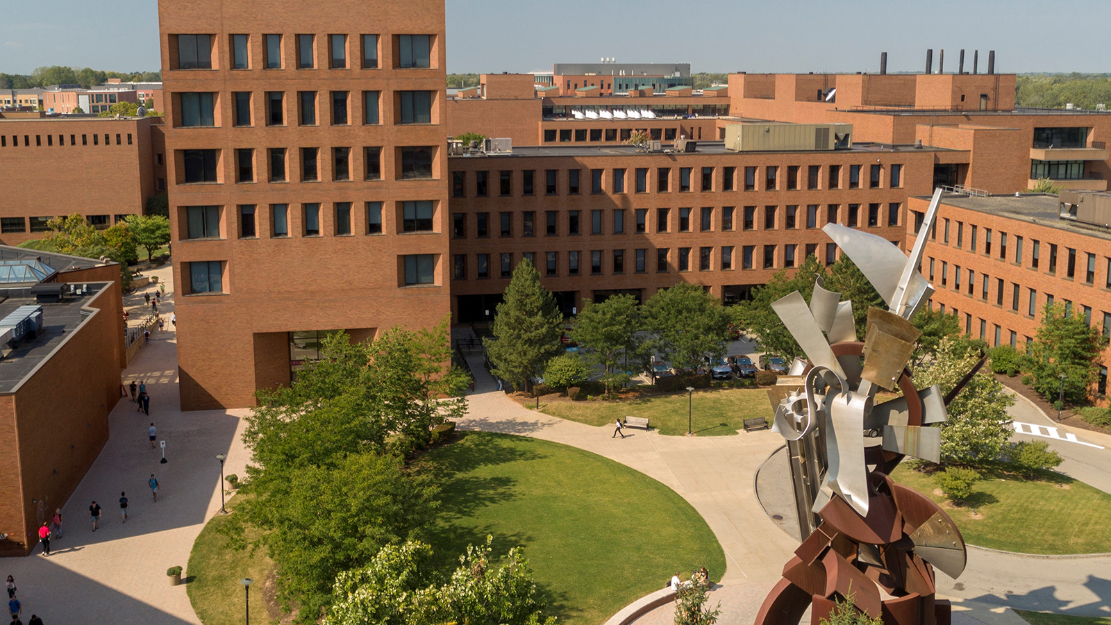 RIT campus panorama with brick buildings, green spaces, walking paths, and an abstract metal sculpture.