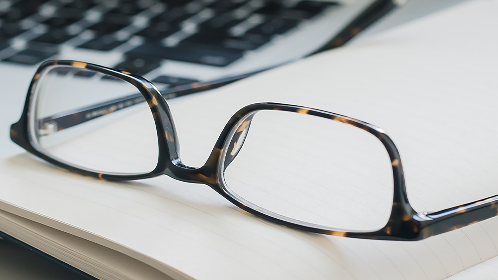 Pair of black-framed eyeglasses on an open notebook next to a laptop keyboard, suggesting a work or study environment.