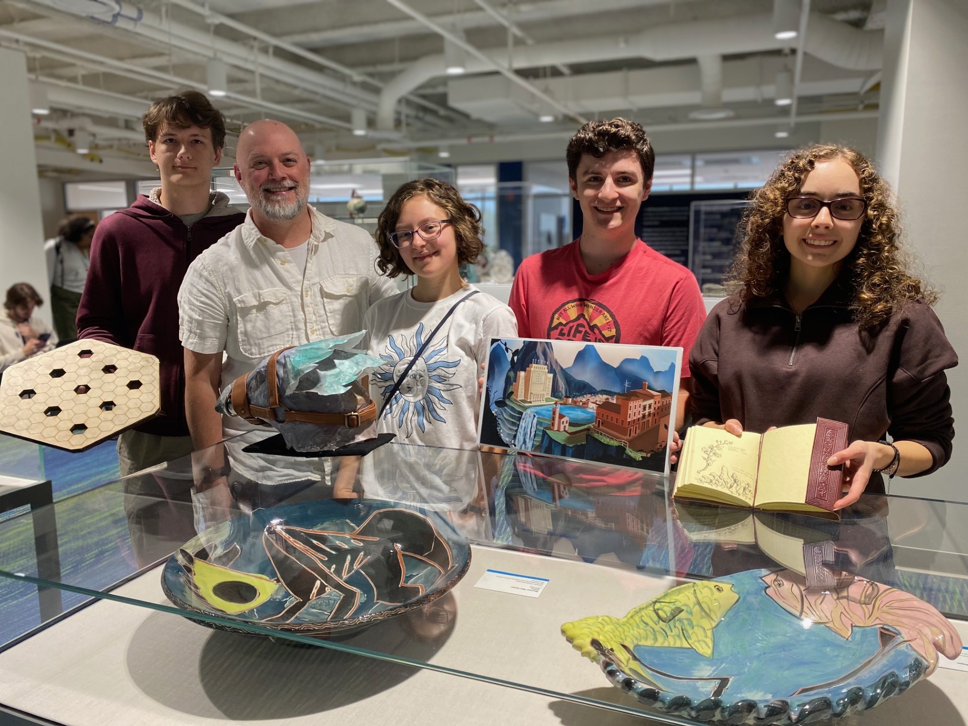 four students with a professor standing behind glass display cases while holding handmade projects.