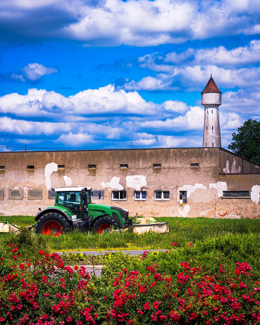 a saturated color photograph off a green tractor in front of a dilapidated masonry building with a watch tower under a brilliant blue sky with puffy clouds.