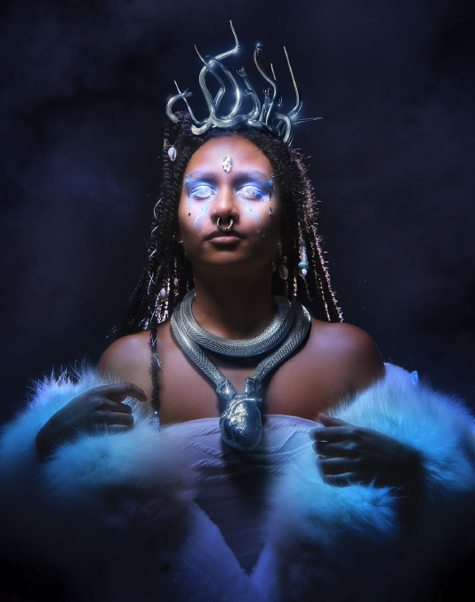 a color photograph of a person on a black background   wearing a light blue   off-the-shoulder gown   a bold serpent necklace and crown with eyes that are whited out and seemingly glowing.