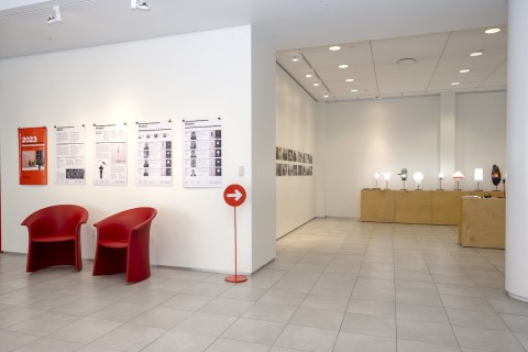 a view into the Grid Space gallery with two red chairs, four information panels and a view of the display of lamps on wood stands. 