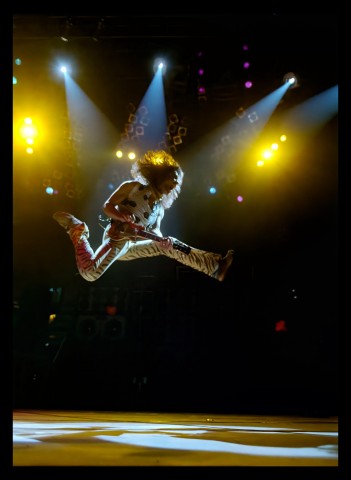 Color photograph of musician Eddie Van Halen playing a guitar on stage while leaping in the air.