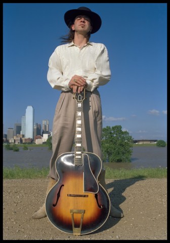 Color photograph of musician Stevie Ray Vaughan standing with his guitar held in front of him.