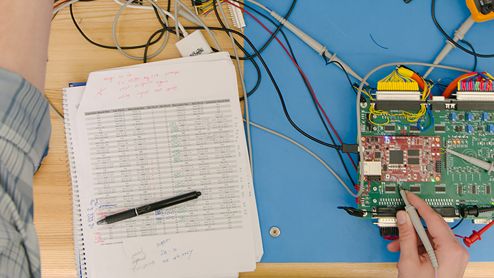 An individual working on an electronic circuit board with a complex wiring setup on a wooden table, alongside a notebook with handwritten notes and mathematical calculations.