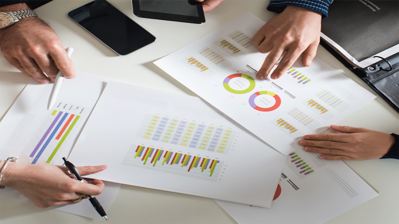 Two individuals analyzing colorful data charts and graphs on white paper sheets during a business meeting, with digital devices nearby on a white table.