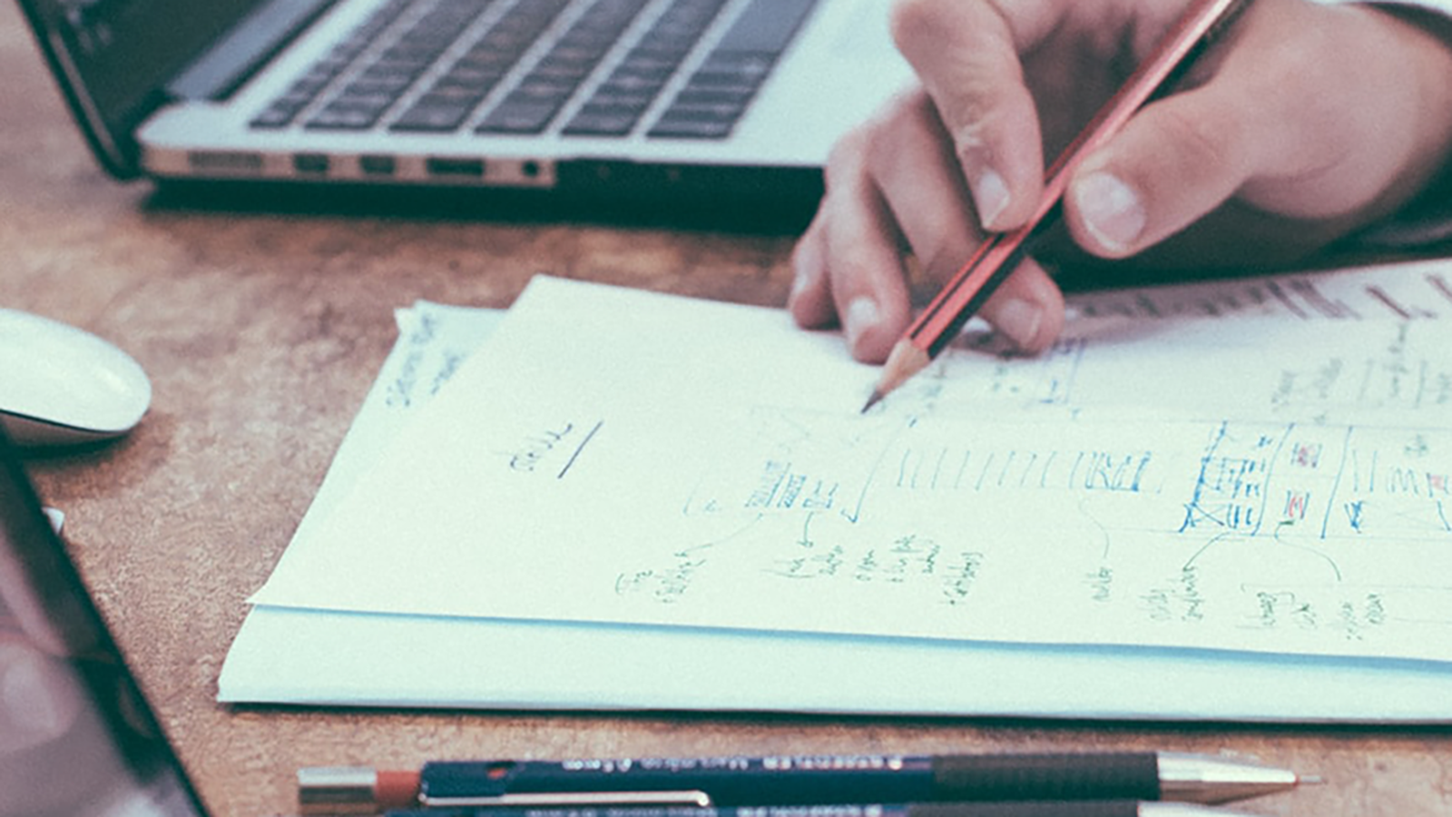 Person writing mathematical equations on a sheet of paper with a pencil, with a laptop and additional stationery items in the background.